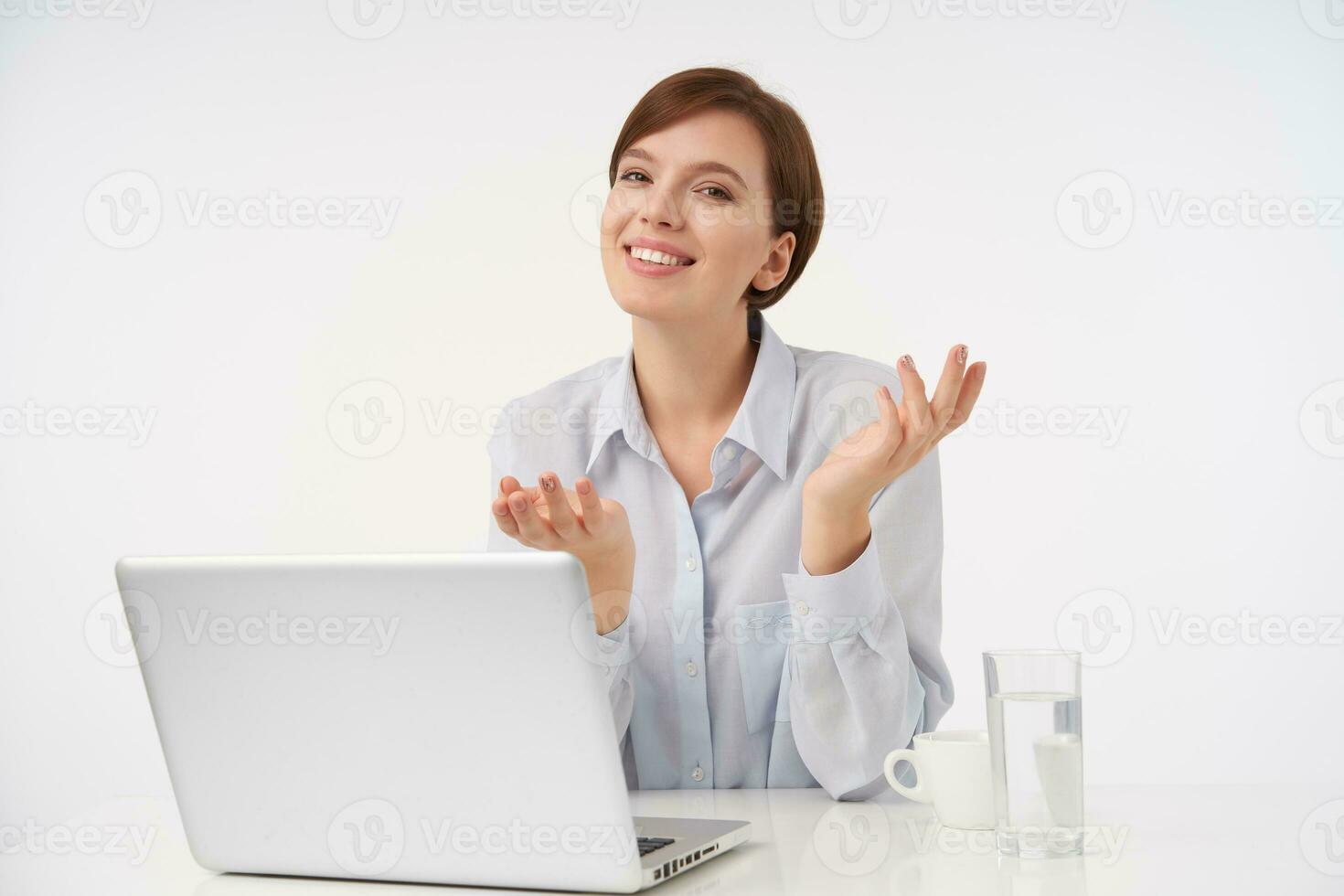 Positive young beautiful brown-eyed short haired lady with natural makeup shrugging with raised palms and smiling positively at camera while sitting over white background in blue shirt photo