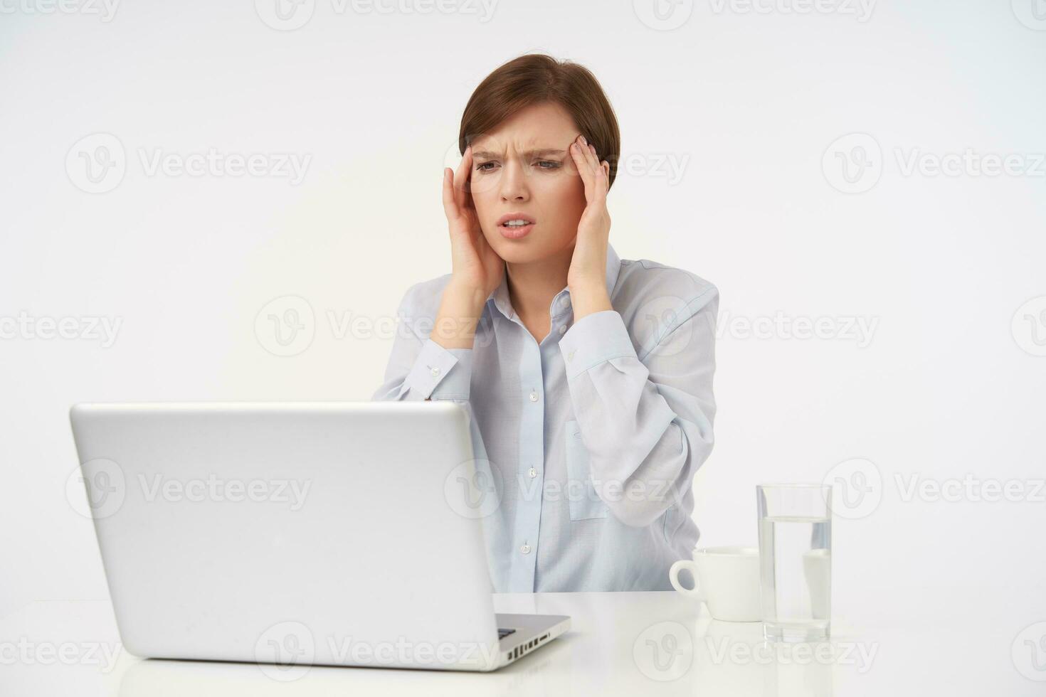 Unhappy young brown-eyed brunette female with casual hairstyle holding hands on temples and frowning face while having heave headache, posing over white background in formal clothes photo