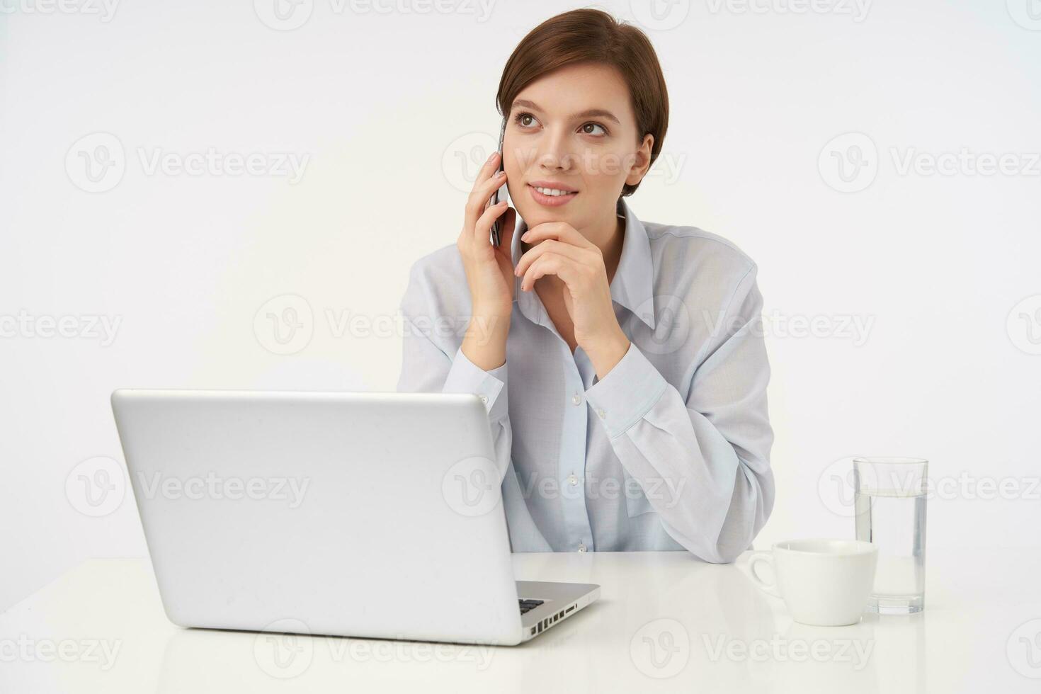 Pleased young attractive brown haired woman with short trendy haircut holding her chin with raised hand and smiling positively while making call, isolated over white background photo