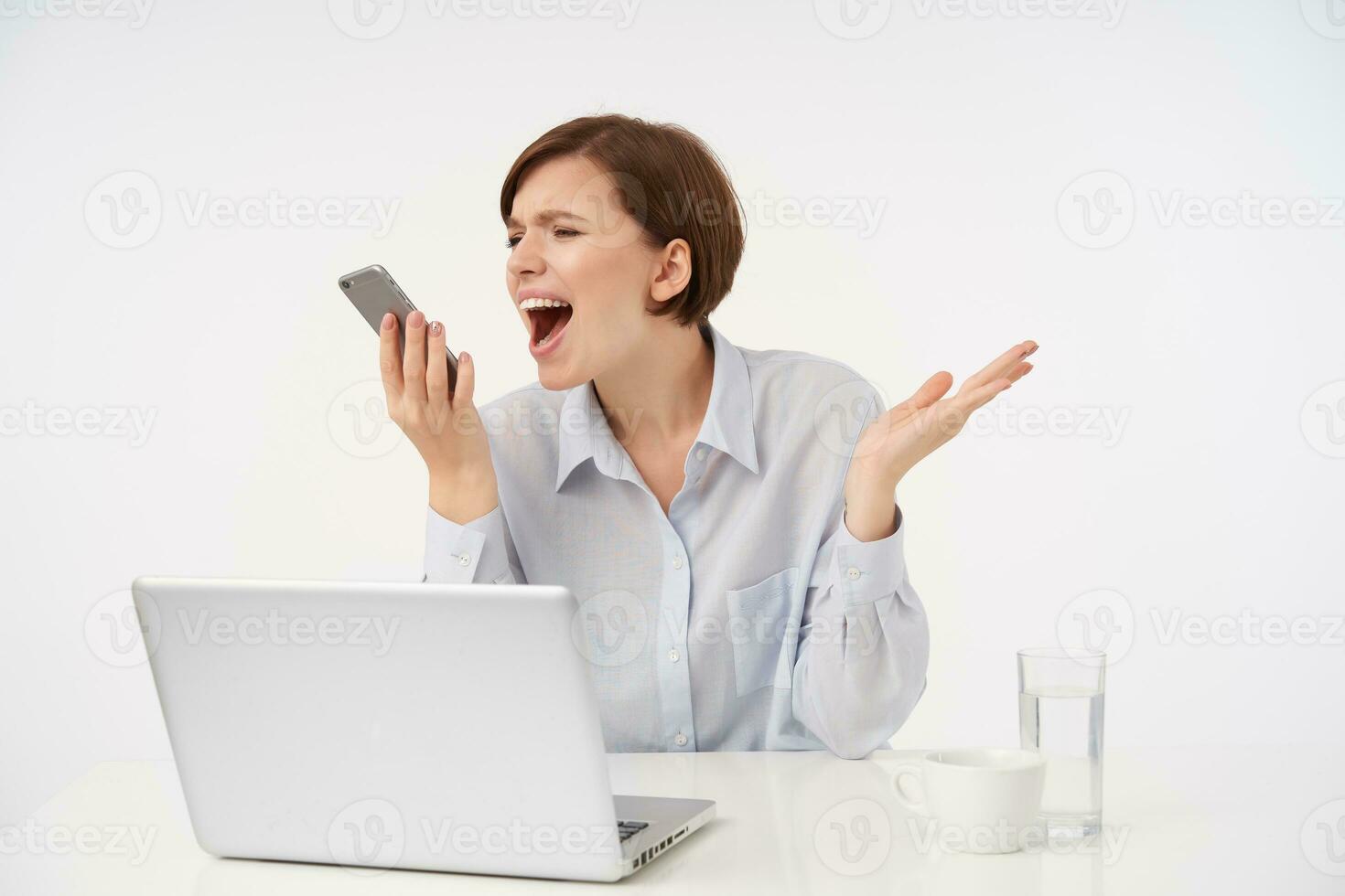 Stressed young brown haired elegant female with short trendy haircut holding handset in raised hand and shouting furiously in it with closed eyes, isolated over white background photo