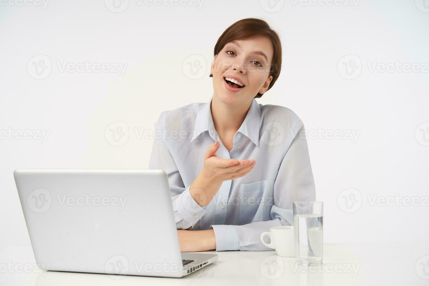 Indoor shot of joyful young brown-eyed short haired lady with casual hairstyle working in modern office with laptop, raising her palm and smiling cheerfully at camera photo