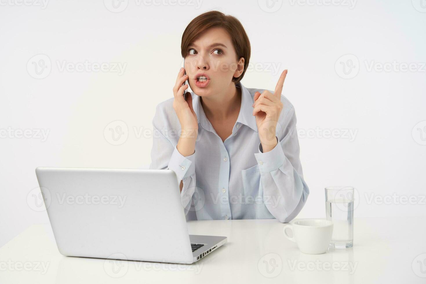 Indoor photo of young attractive brunette lady with short trendy haircut keeping her forefinger raised while having tense phone conversation, wearing blue shirt while posing over white background