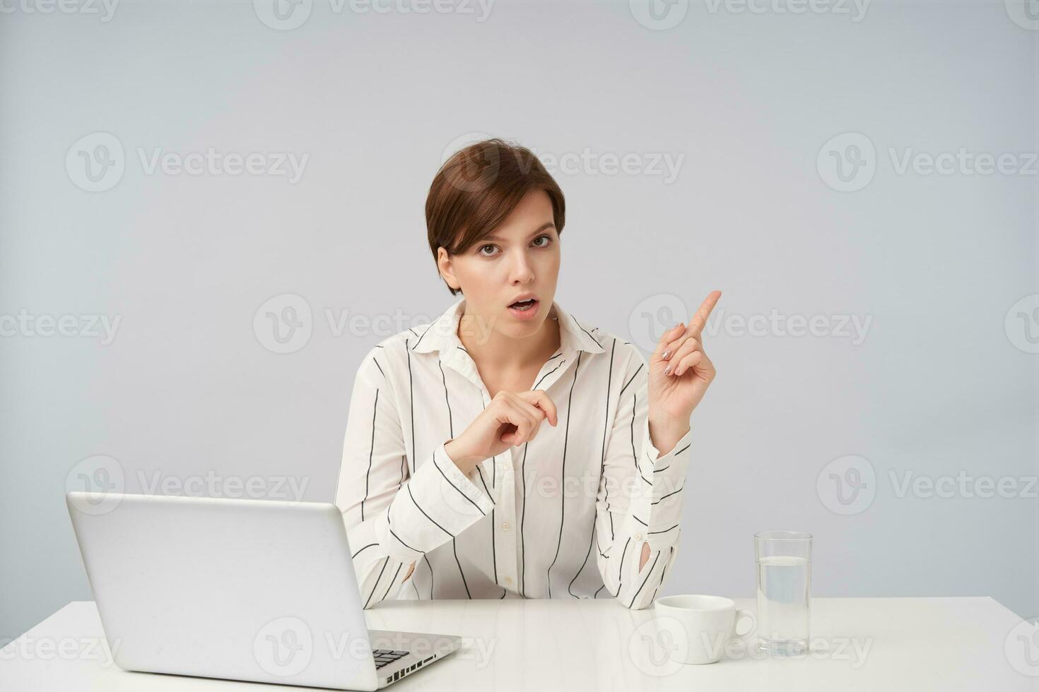 Confused young attractive brown-eyed short haired lady with natural makeup pointing aside with forefingers and looking at camera with puzzled face, isolated over white background photo