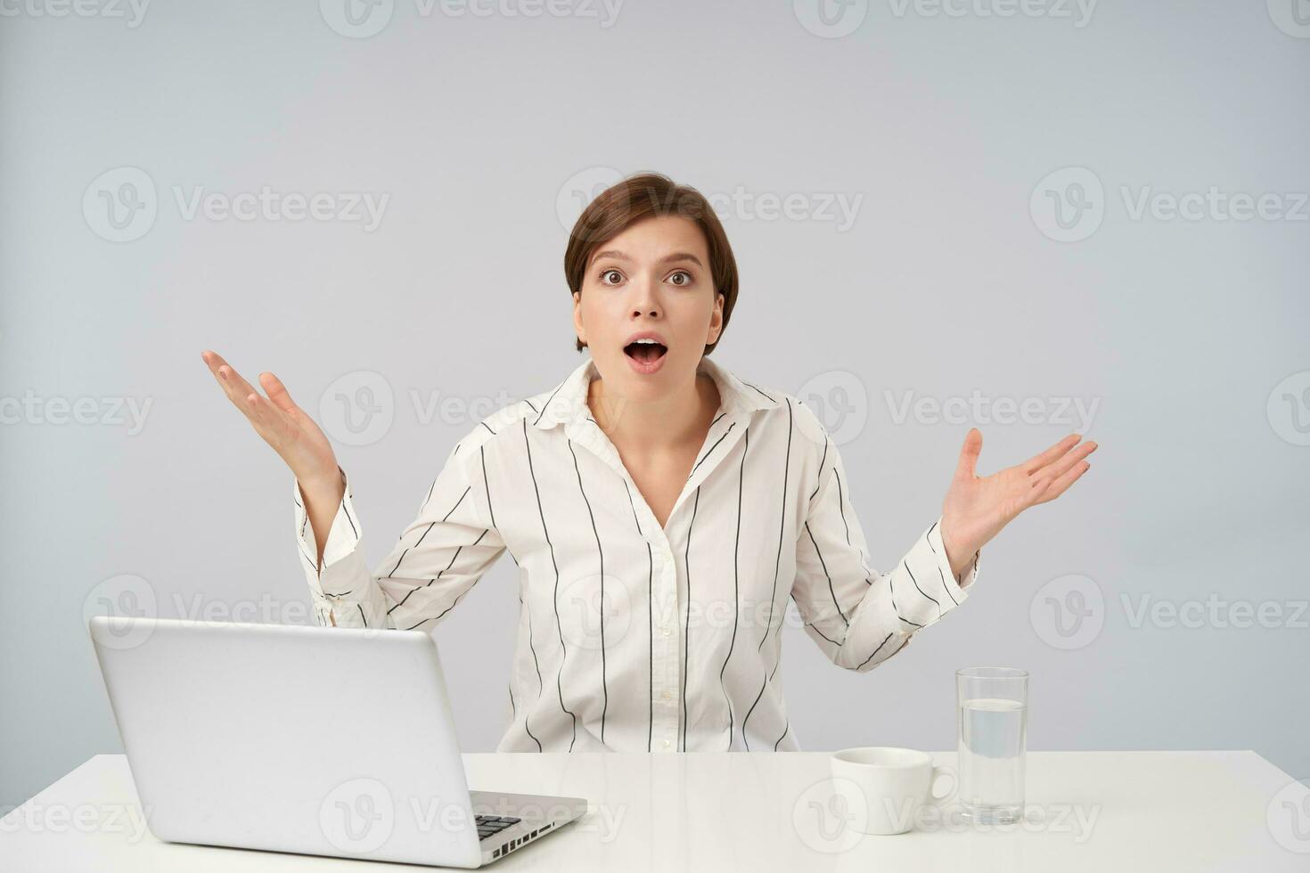 Agitated young brown haired woman with short trendy haircut looking at camera with wide eyes and mouth opened and raising excitedly palms, isolated over white background photo
