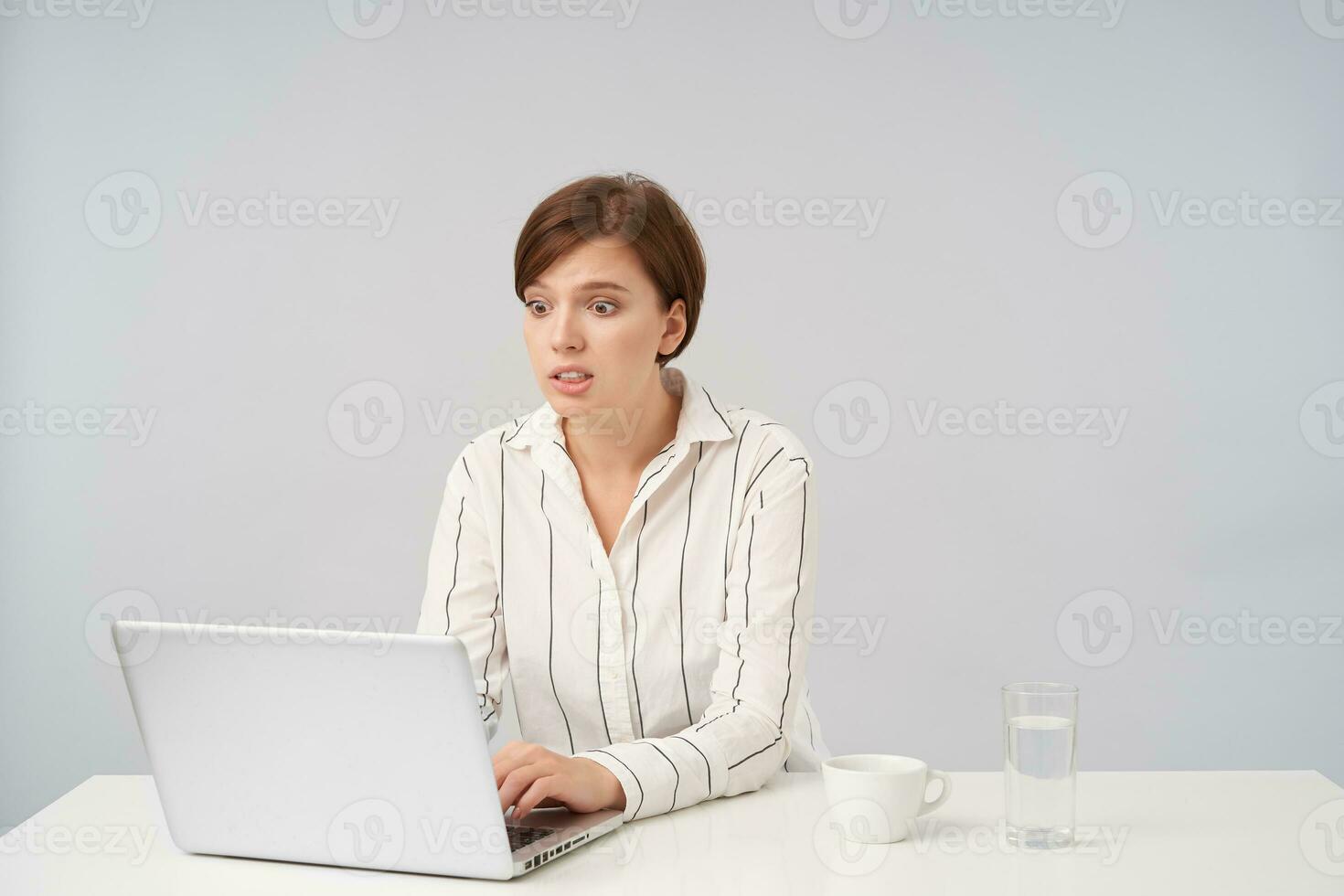 Agitated young attractive short haired brunette female looking confusedly on her laptop while typing text on keyboard, dressed in striped shirt while sitting over white background photo