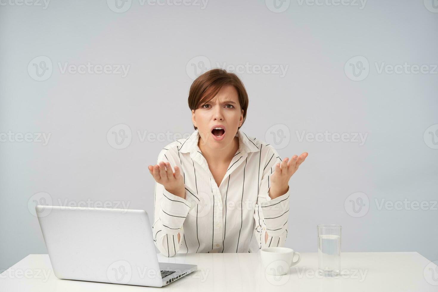 Perplexed young brown haired woman with short trendy haircut raising excitedly hands while frowning her eyebrows with opened mouth, sitting over white background photo