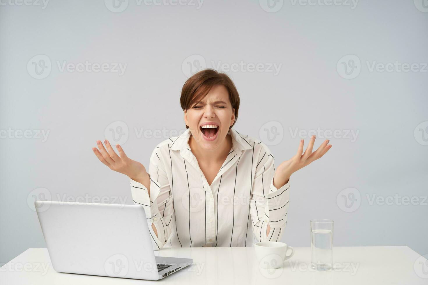Stressed young short haired brunette woman with natural makeup keeping her eyes closed while shouting heatedly with raised hands, wearing striped shirt over white background photo