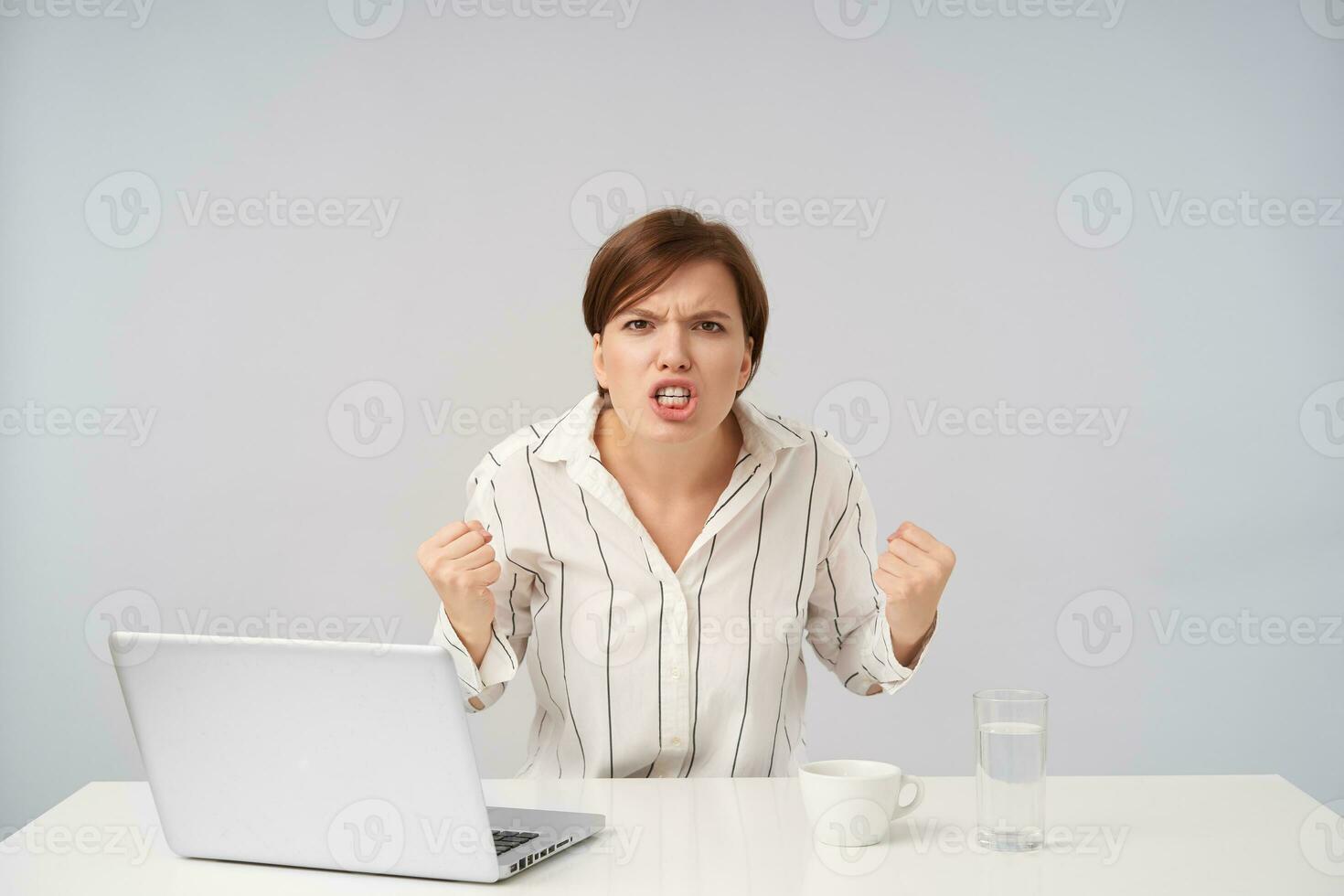 Furious young brown haired female with short trendy haircut clenching her fists and frowning face while looking violently at camera, posing over white background in striped shirt photo