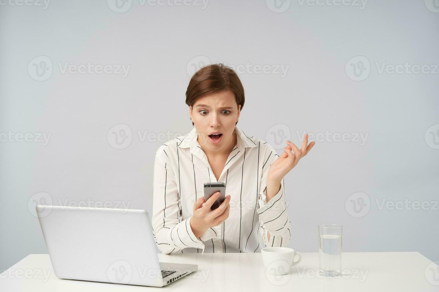 Puzzled young brown haired female with short haircut holding mobile phone and looking confusedly at screen, raising perplexedly palm while sitting over white background photo
