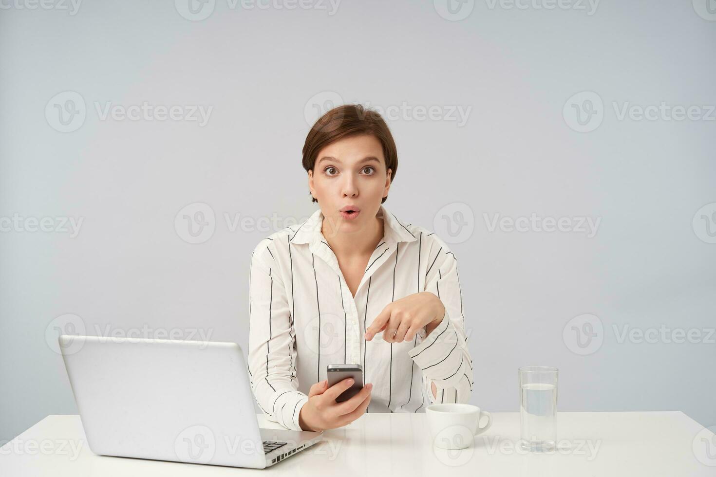 Surprised young lovely short haired brunette lady dressed in striped shirt showing on smartphone in her hand and rounding her brown eyes while looking amazedly at camera, sitting over white background photo
