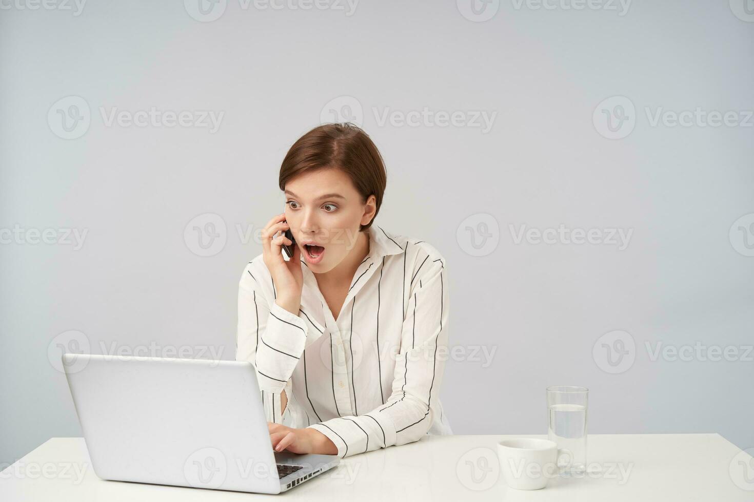 Amazed young pretty short haired brunette lady working with her laptop and looking surprisedly on screen during phone conversation, sitting at table over white background photo