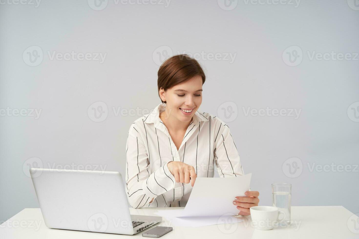 Cheerful young pretty brown haired lady in formal clothes sitting at table over white background, reading text on piece of paper and being pleased with it photo