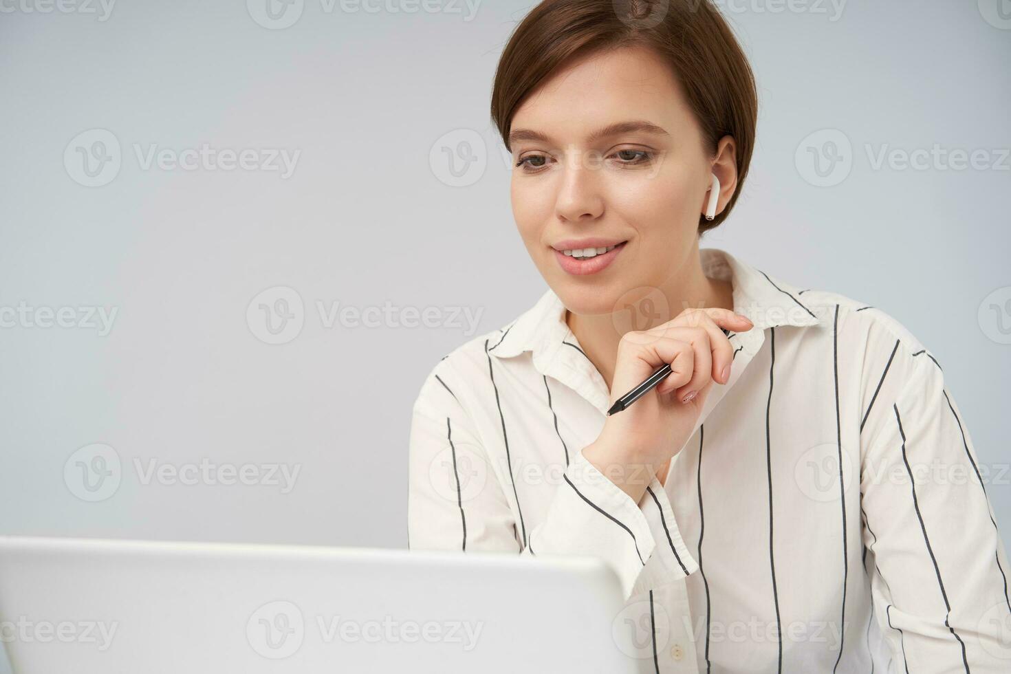 Portrait of beautiful young short haired brunette lady with trendy haircut raising hand with pen in it and smiling positively while looking on screen of her laptop, posing over white background photo