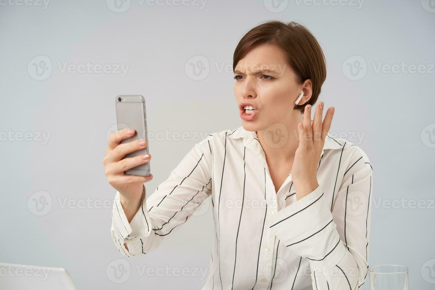 disgustado joven bonito corto peludo morena hembra ceñudo su Cejas y mirando a pantalla con abadejo mientras teniendo Estresante teléfono conversación, posando terminado blanco antecedentes foto