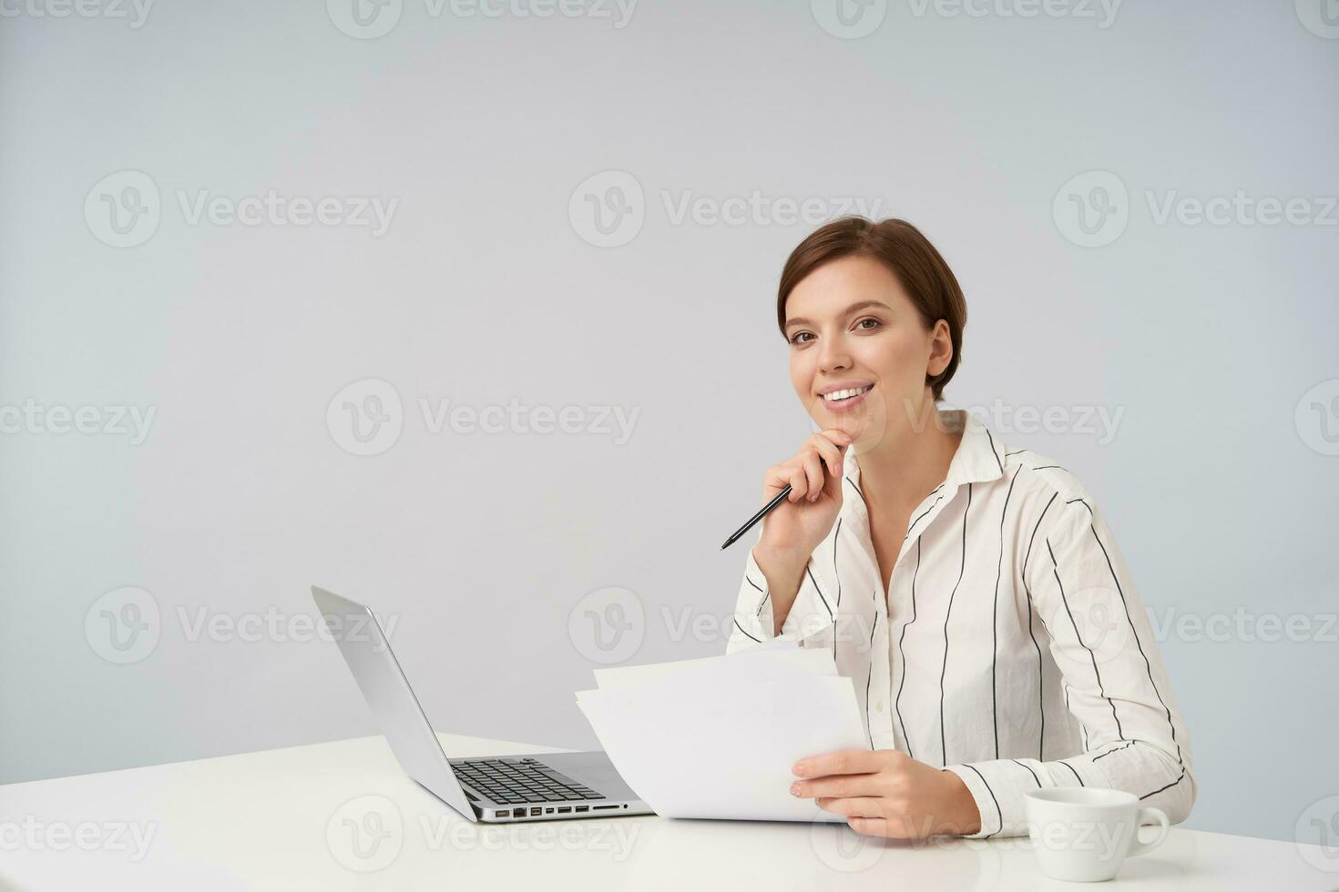 Cheerful young attractive brown-eyed short haired woman smiling pleasantly while looking at camera and leaning chin on raised hand, posing over white background with laptop and documents photo