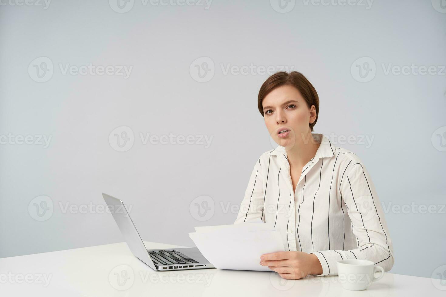 Bewildered young pretty short haired brunette lady with natural makeup sitting at table over white background and holding piece of paper, squinting while looking at camera with serious face photo