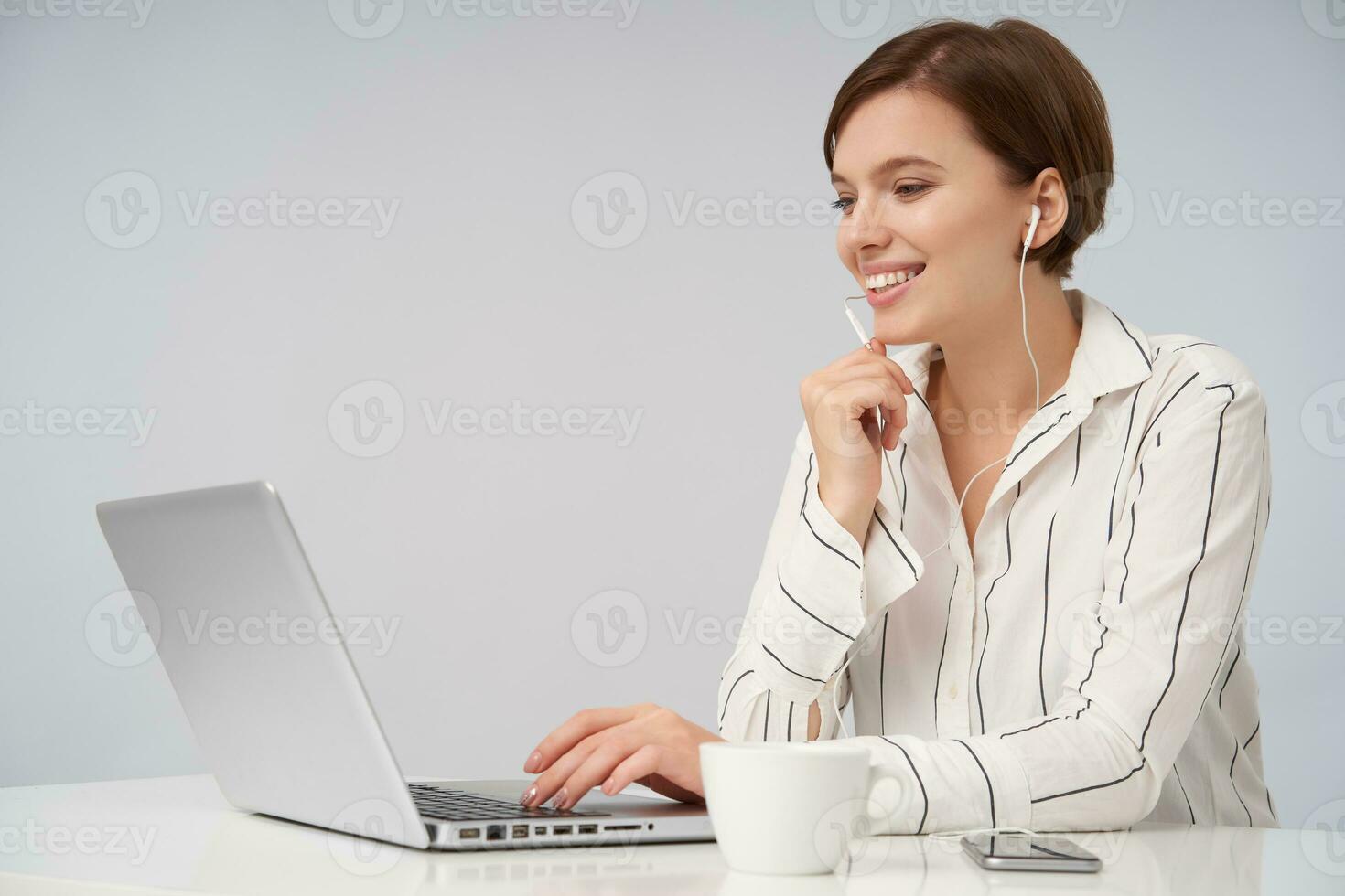 Shot of attractive young brown haired woman with short trendy haircut holding hand on keyboard while sitting over pink background with laptop, smiling happily and holding raised hand under her chin photo