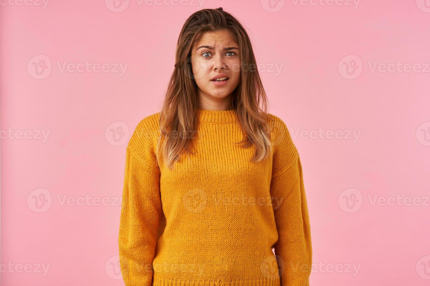 Puzzled young attractive brown haired woman raising wonderingly eyebrow and wrinkling her forehead while posing over pink background in warm knitted mustard sweater photo