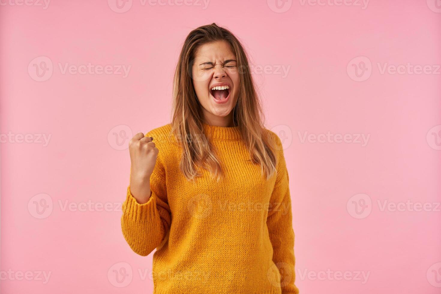 Indoor photo of good looking young brunette lady frowning her face with closed eyes while screaming loud, raising up her fist while standing over pink background