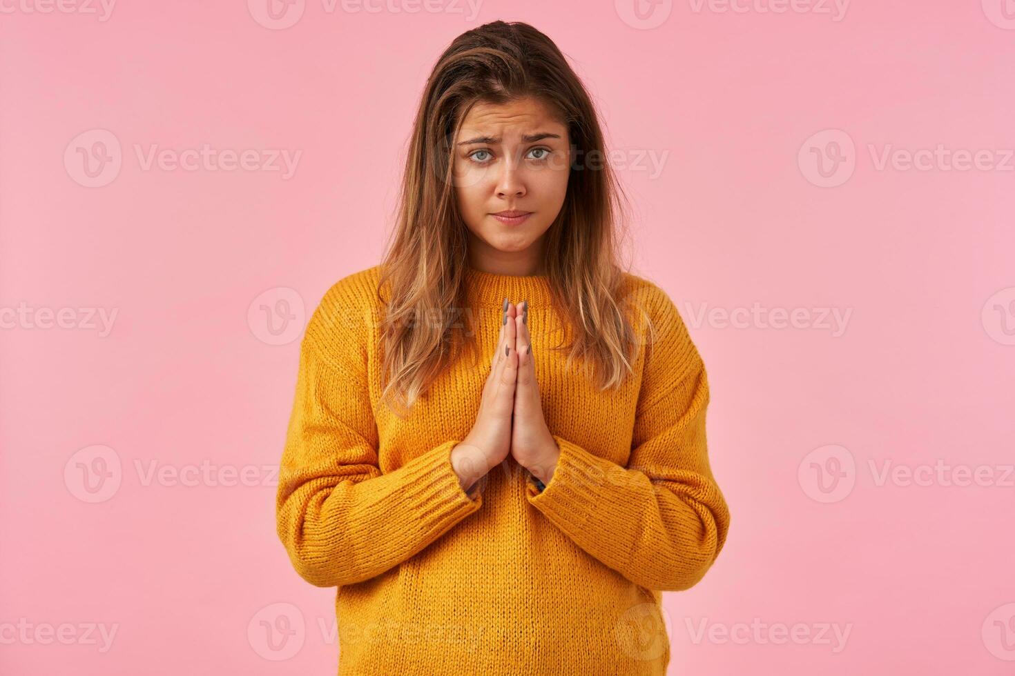 Desperate young pretty brown haired woman looking sadly at camera with folded lips, raising palms in praying gesture while standing against pink background photo