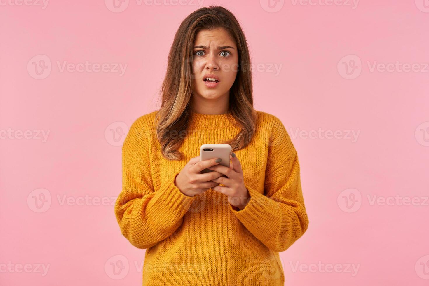 Puzzled young pretty brown haired lady with casual hairstyle holding mobile phone and looking at camera with confused face, dressed in mustard knitted sweater over pink background photo