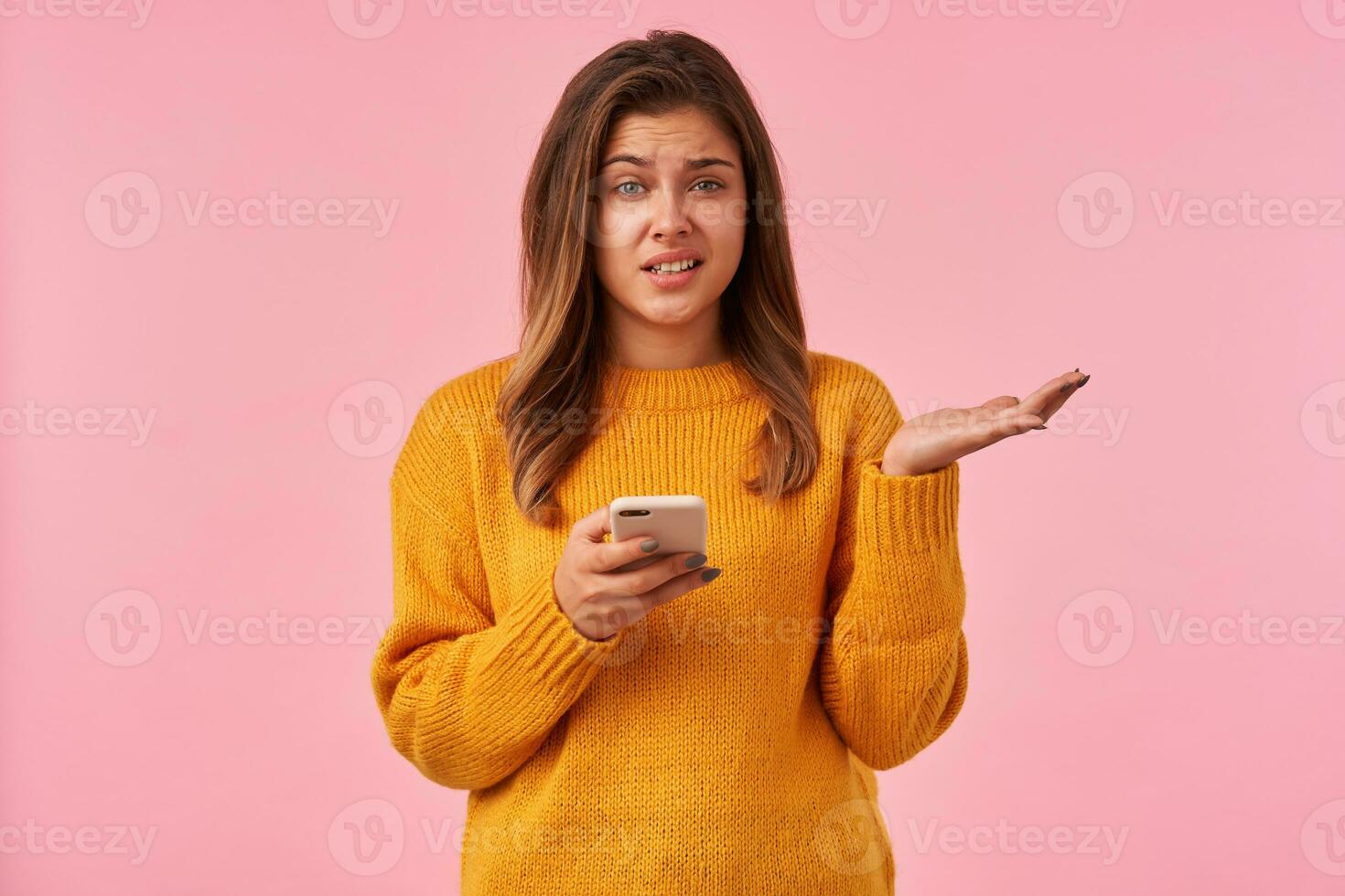 Indoor shot of young brown haired confused female looking at camera with displeased face and raising confusedly hand while standing against pink background with smartphone photo
