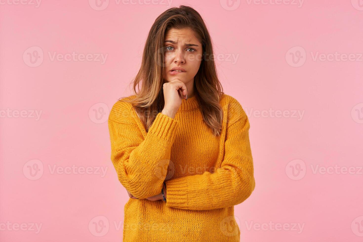 Confused young green-eyed brunette female keeping hand on her chin and raising eyebrow while looking at camera, biting underlip while posing over pink background photo