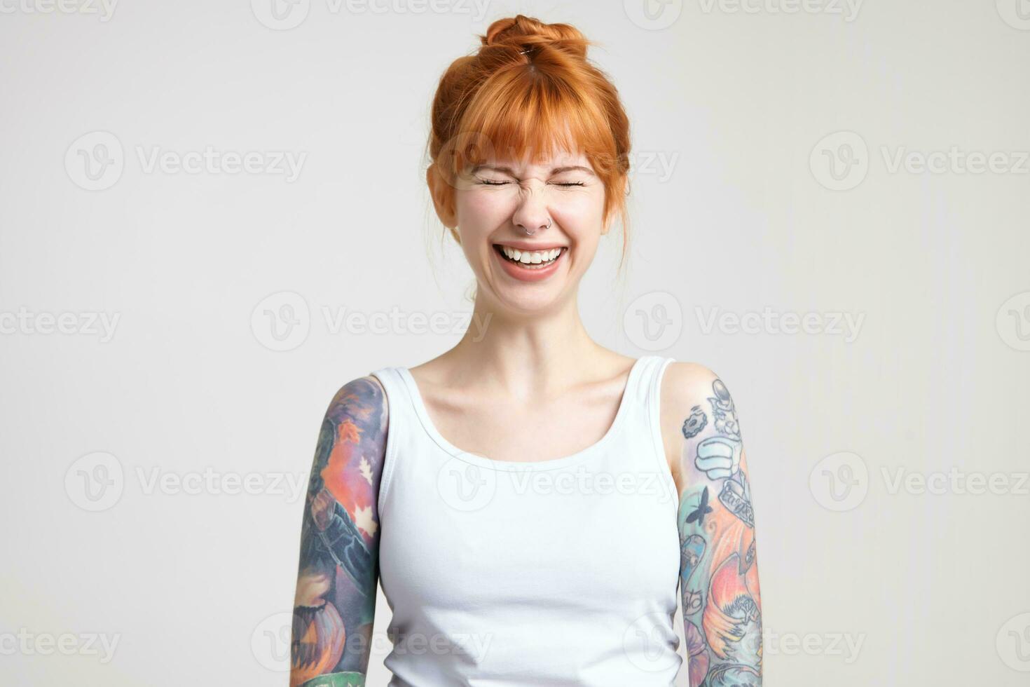Joyful young attractive redhead woman with tattoos keeping her eyes closed while laughing cheerfully, dressed in white shirt while posing over white background photo