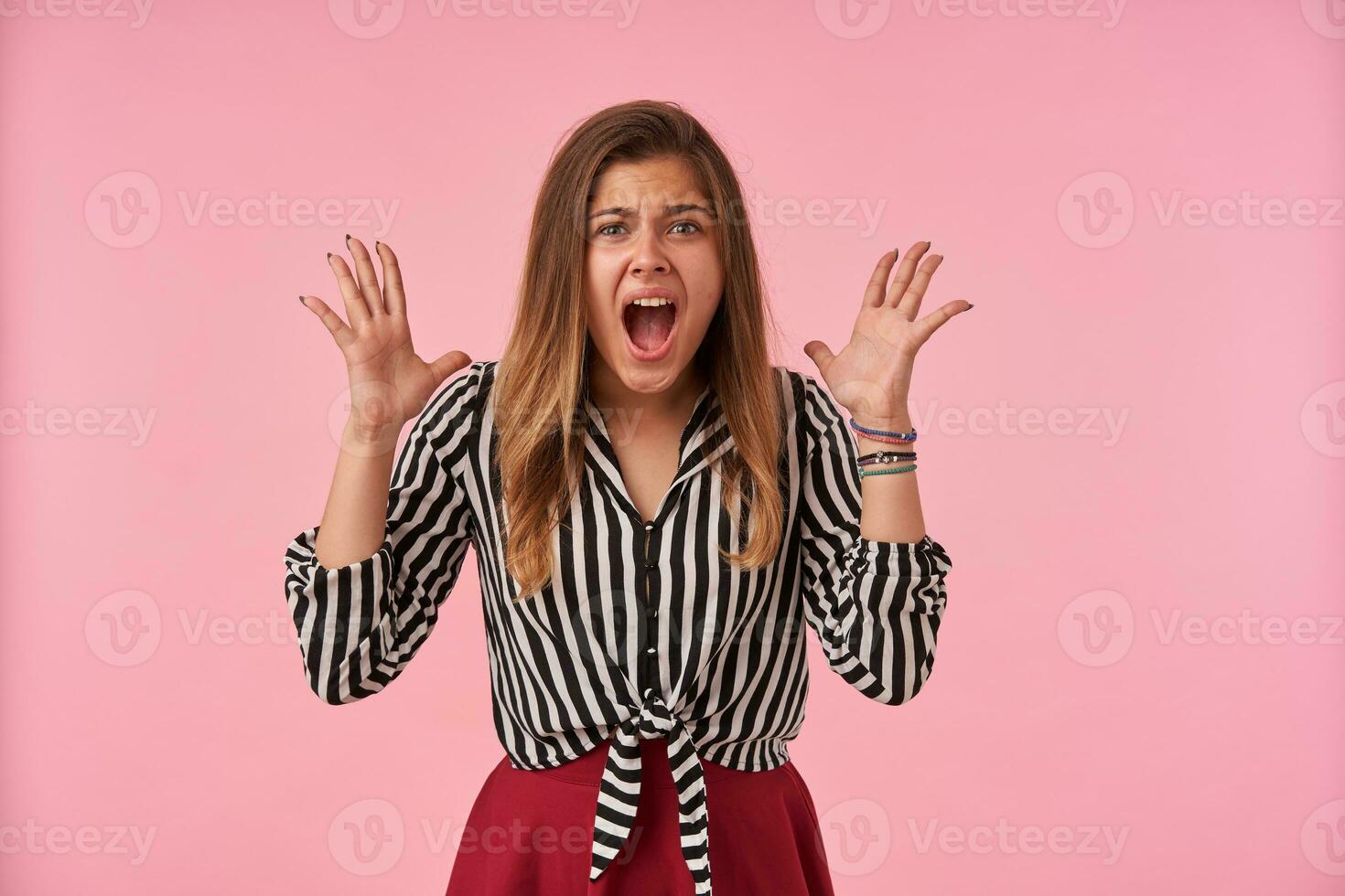 Agitated young attractive brunette woman with casual hairstyle shouting loud and raising emotionally her hands while standing over pink background in festive clothes photo