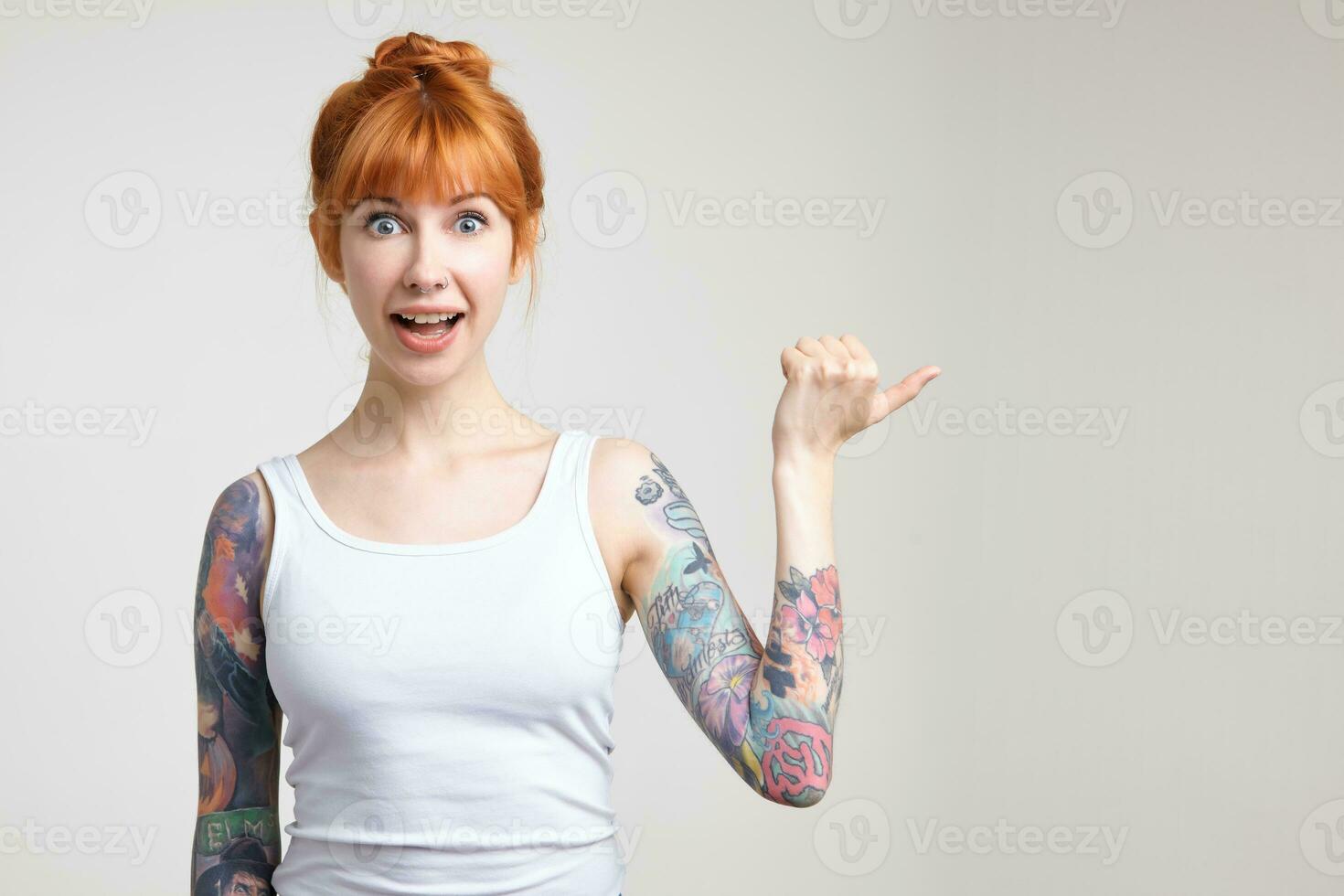 Excited young attractive redhead tattooed female rounding surprisedly her eyes while showing aside with raised hand, isolated over white background in casual shirt photo