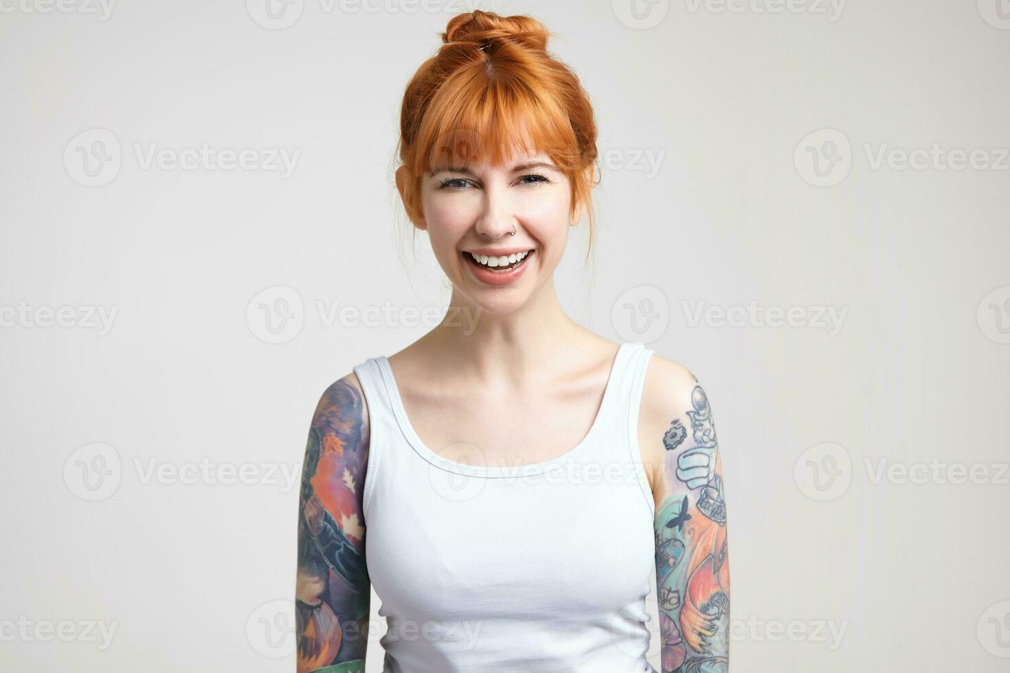 Joyful young attractive tattooed woman with foxy hair laughing happily while looking cheerfully at camera, standing over white background with hands down photo