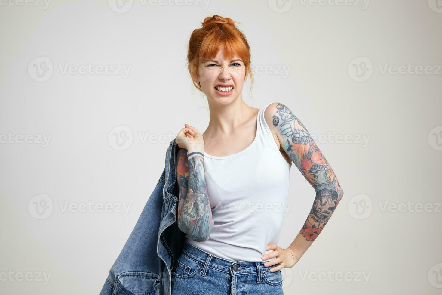 Horizontal shot of young pretty redhead lady with tattoos holding jeans coat with raised hand and frowning her face while looking at camera, isolated over white background photo