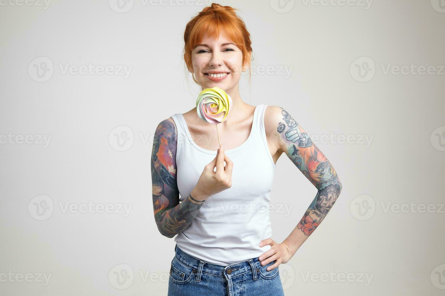 Studio shot of young happy redhead tattooed lady looking cheerfully at camera with wide smile while holding candy on stick, isolated over white background photo
