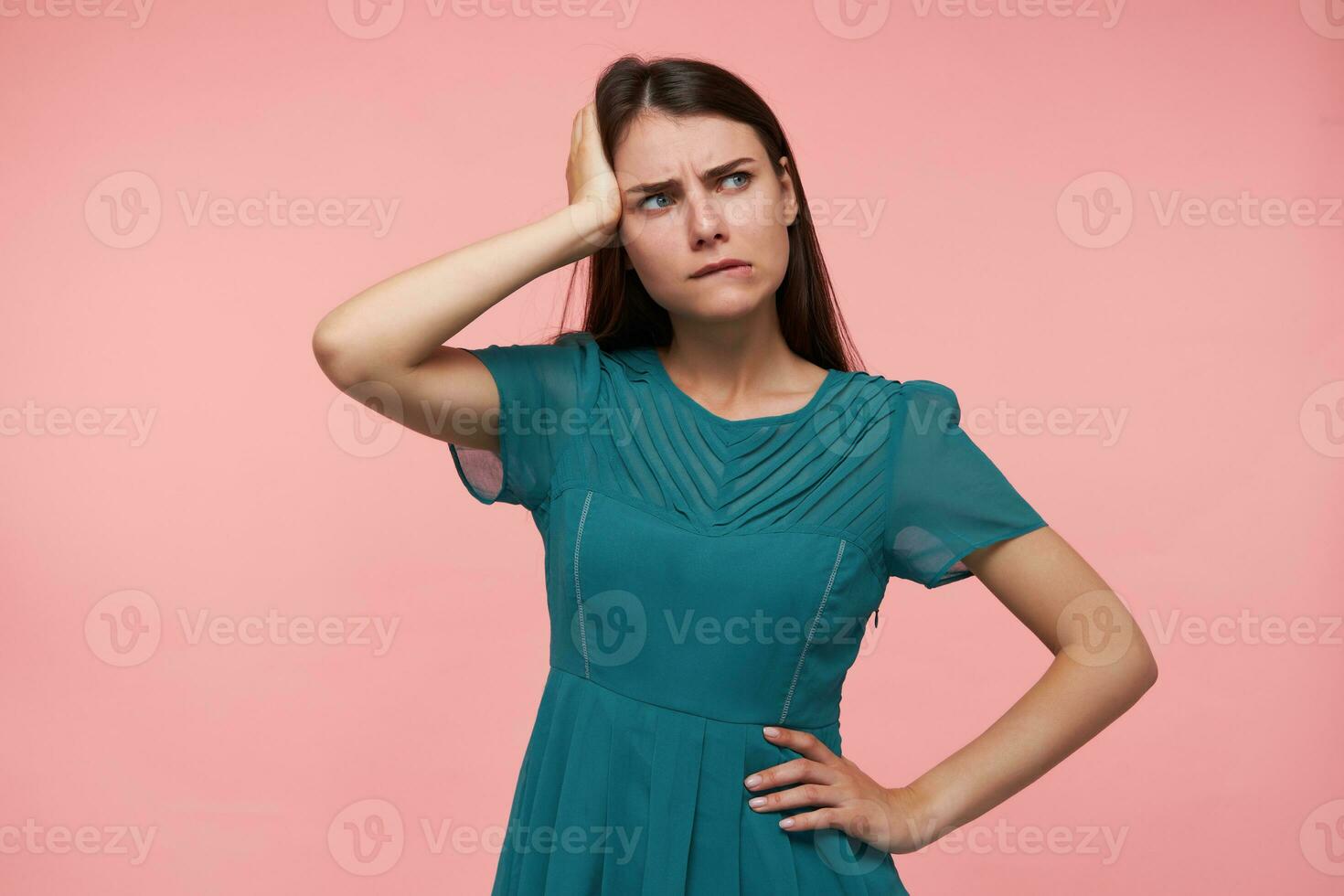 Young lady, pretty woman with long brunette hair. Holding one hand on a waist and touching her head, thinking. Watching to the right at copy space over pastel pink background. Wearing emerald dress photo