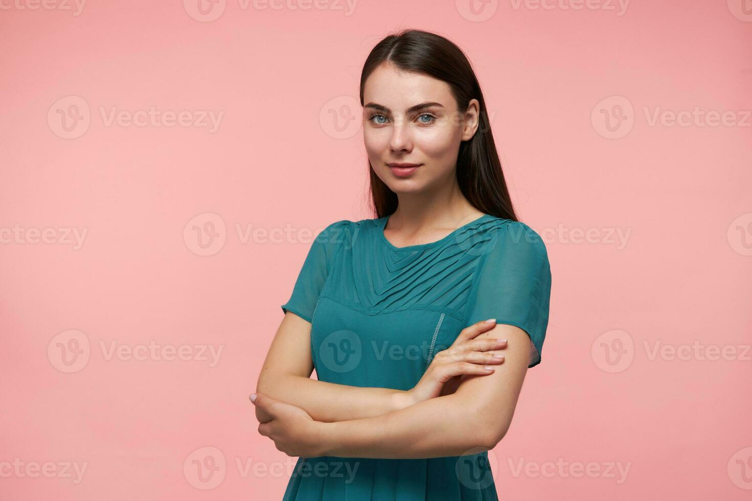 Young lady, pretty woman with long brunette hair. Folding hands on a chest. Wearing emerald dress. Watching at the camera and smile isolated over pastel pink background photo