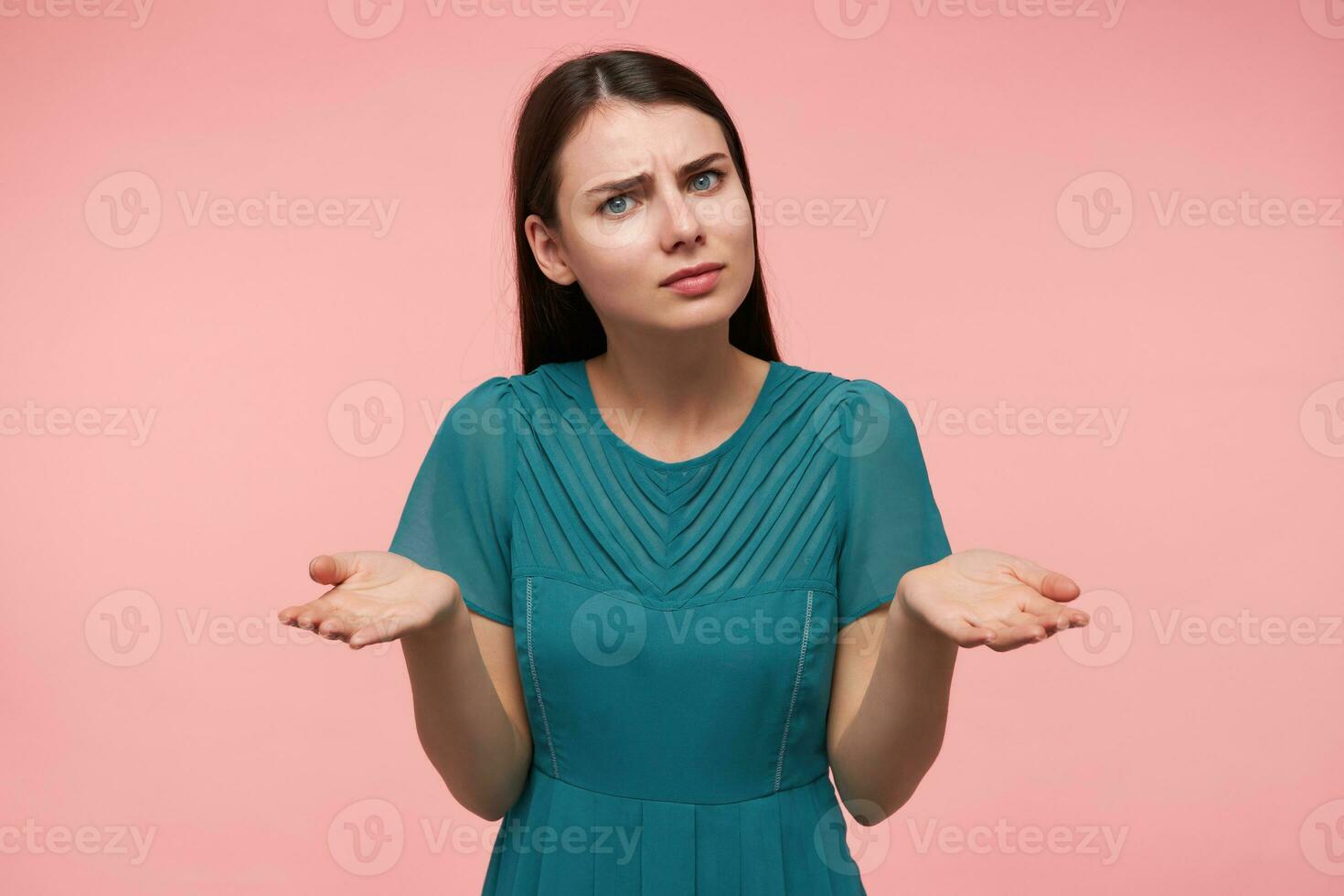 Portrait of attractive, nice looking girl with long brunette hair. Watching at the camera and holding her hands with palms up, copy space, isolated over pastel pink background. Wearing emerald dress photo