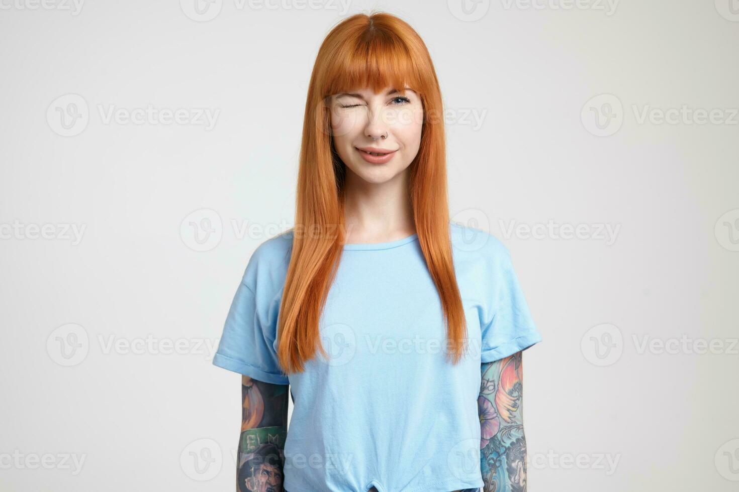 Joyful young attractive tattooed female with loose foxy hair giving positively wink at camera while standing over white background with hands along her body photo