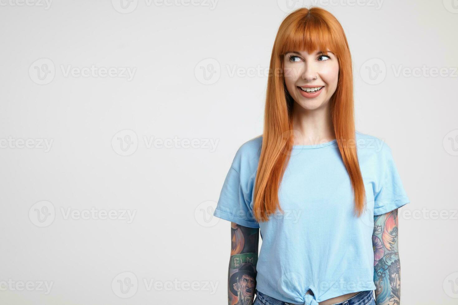 alegre joven atractivo largo peludo pelirrojo dama con casual peinado sonriente extensamente mientras mirando con alegría aparte, posando terminado blanco antecedentes en azul camiseta foto