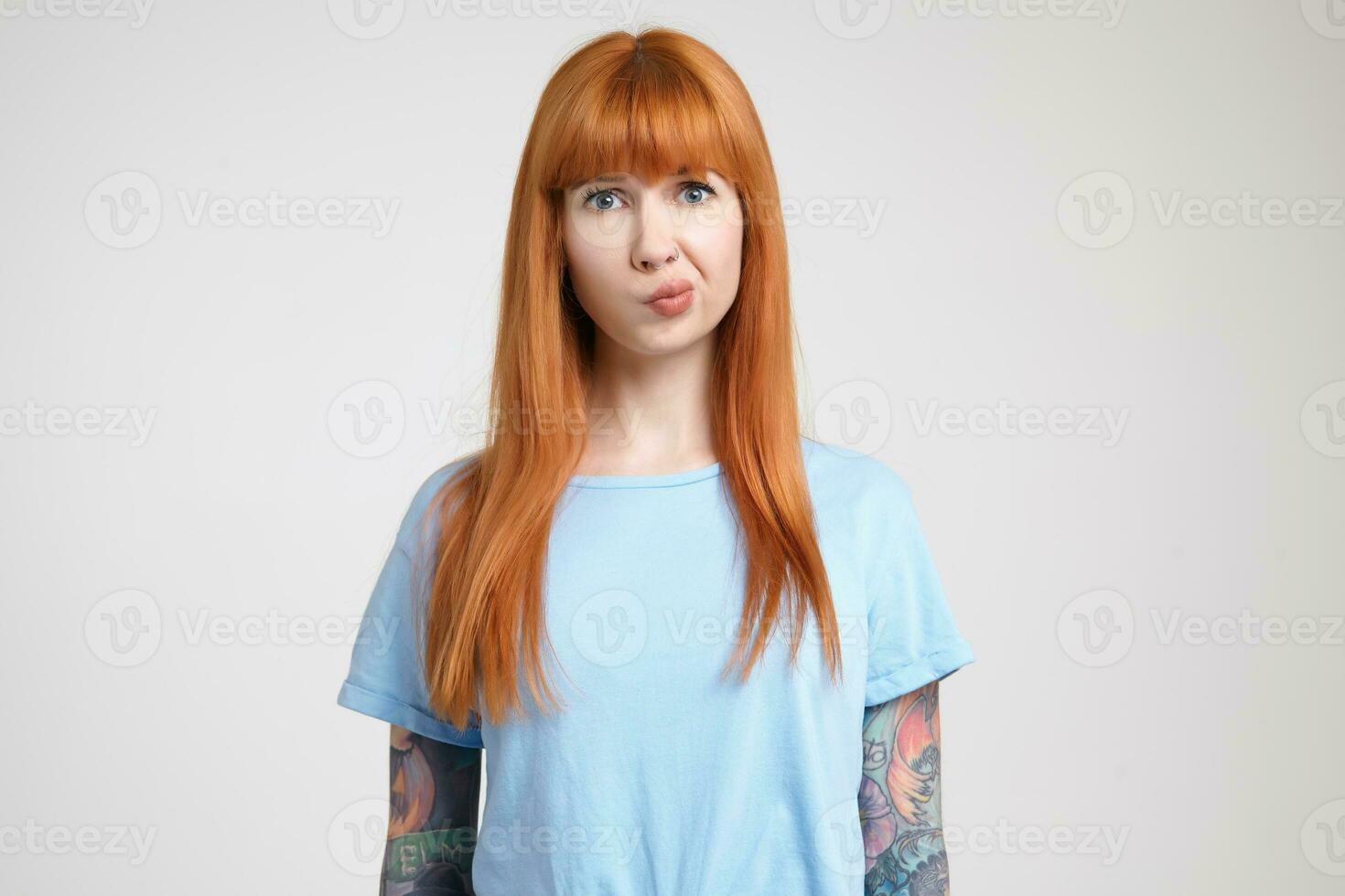 Indoor shot of confused young tattooed female with loose foxy hair pursing her lips while looking pensively at camera, standing over white background photo