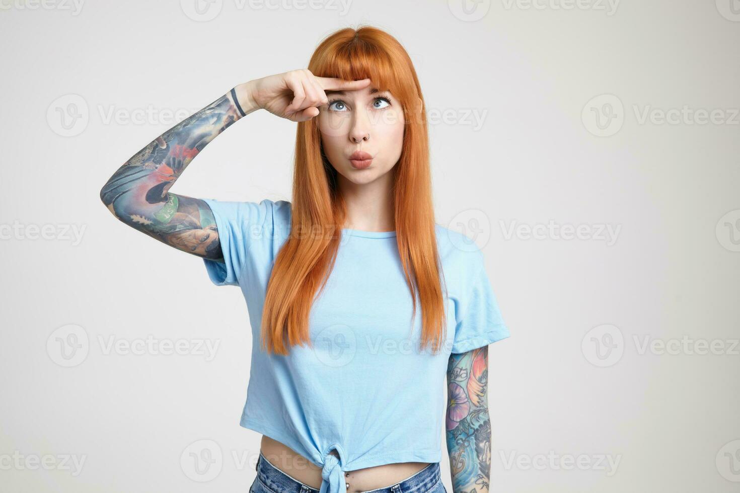 Studio shot of young long haired redhead lady with nose piercing pouting her lips while looking confusedly on her raised forefinger, isolated over white background photo