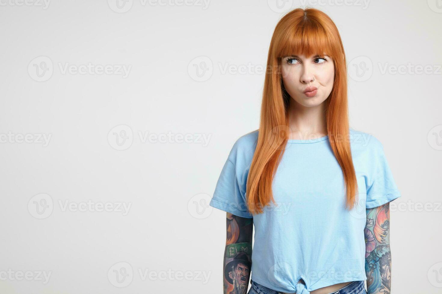 Pensive young pretty redhead tattooed female looking severely aside and twisting her mouth while standing against white background in blue t-shirt photo