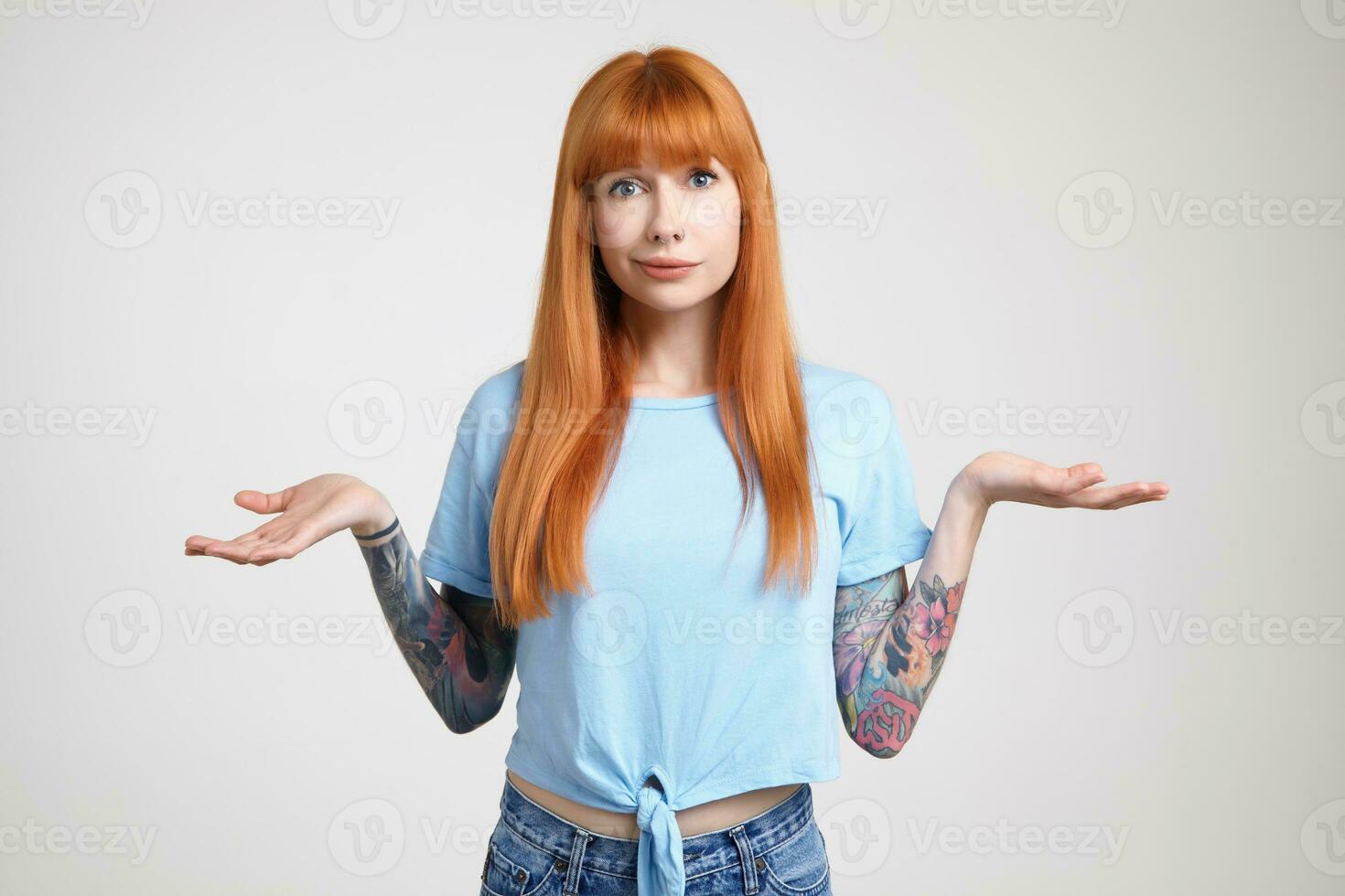 Studio photo of young long haired redhead lady with tattoos keeping her palms raised while looking confusedly at camera, isolated over white background