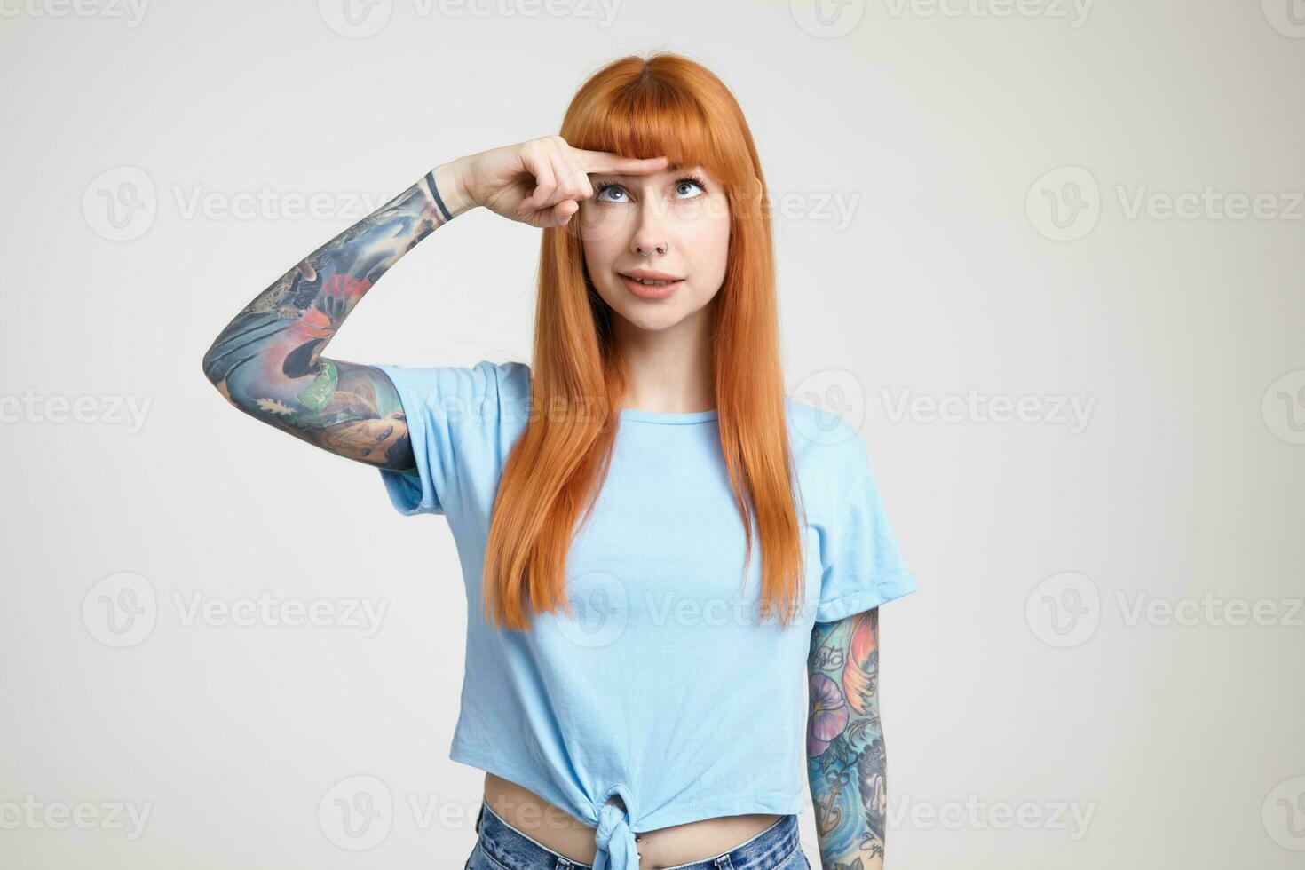 Bewildered young pretty tattooed woman with loose long hair keeping index finger on forehead and looking at it while standing over white background in blue t-shirt photo