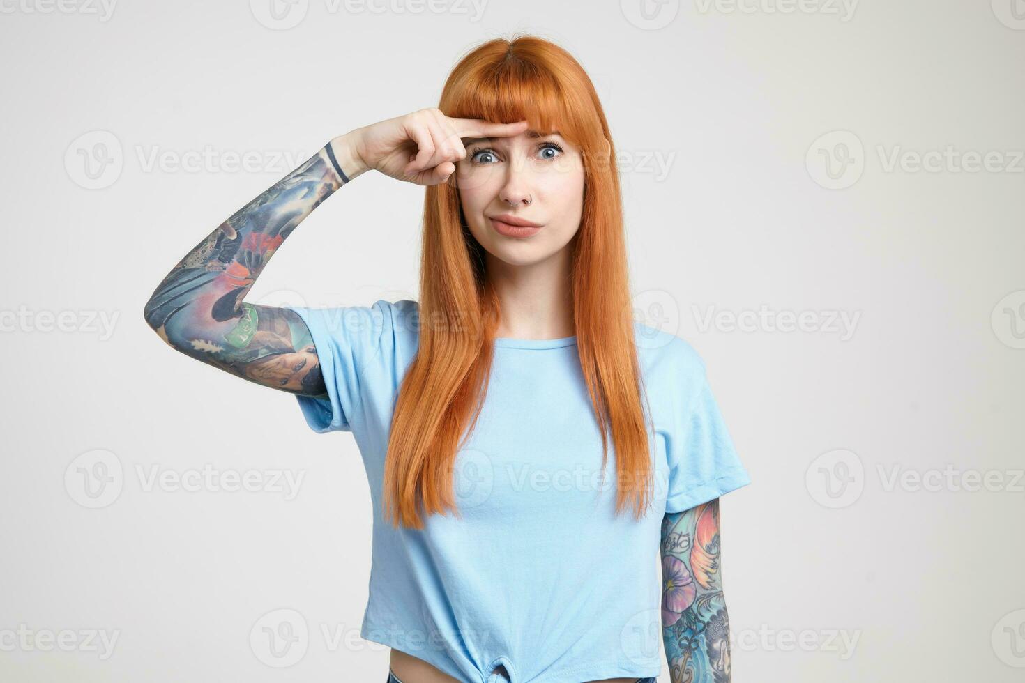 Indoor photo of young long haired redhead woman with tattoos keeping forefinger above her eyebrows and grimacing her face while looking at camera, isolated over white background