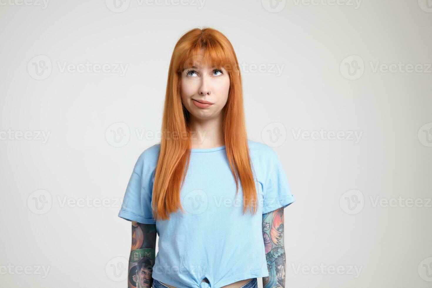 Puzzled young lovely redhead tattooed woman with casual hairstyle blowing on her hair while looking upwards, standing over white background with hands down photo