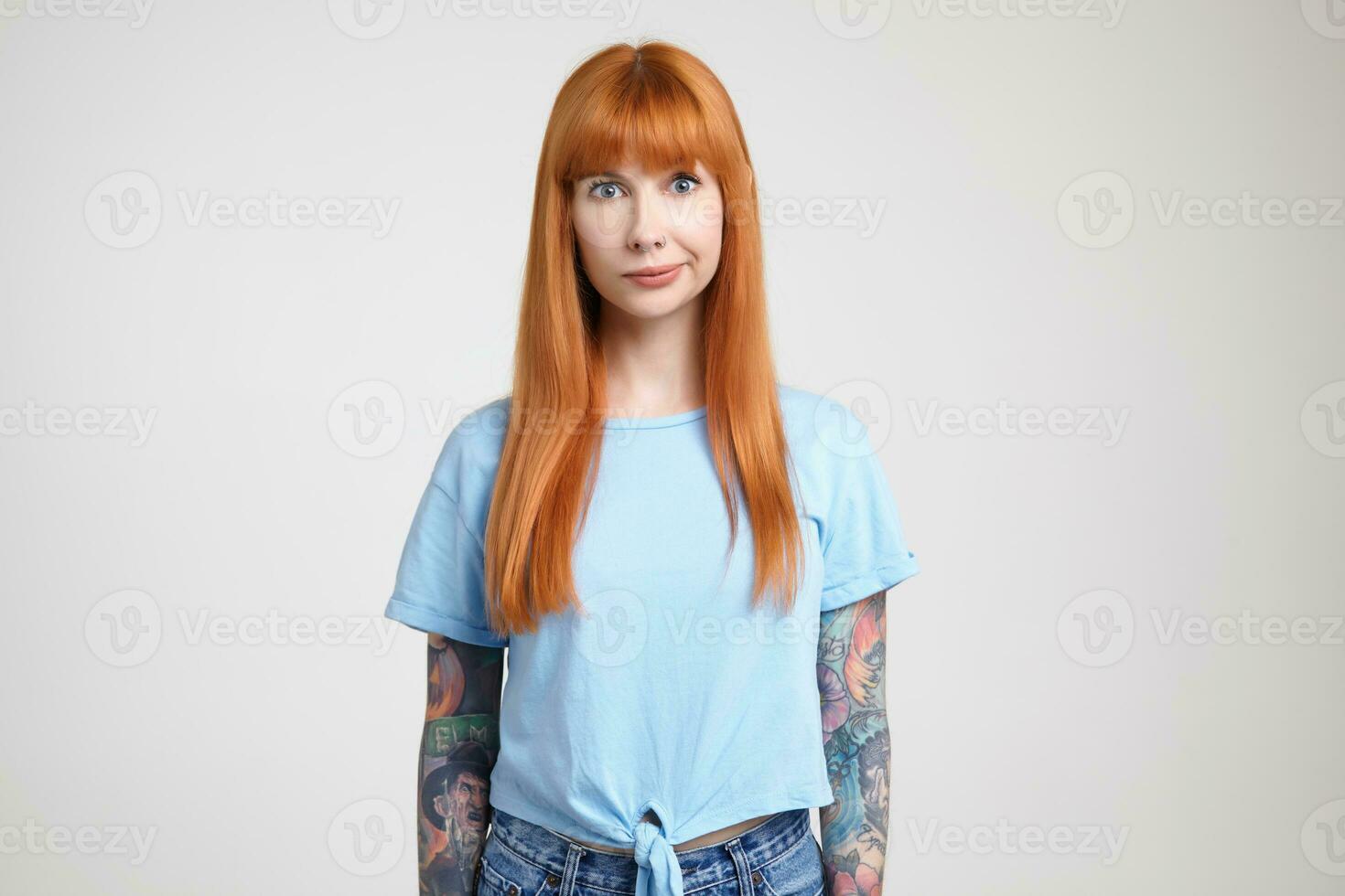 Puzzled young pretty green-eyed long haired woman with loose foxy hair folding her lips while looking confusedly at camera, standing over white background photo