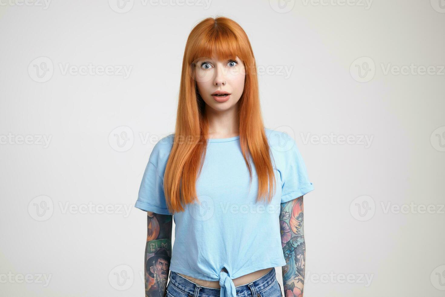 Dazed young attractive long haired redhead lady with tattoos rounding surprisedly her green eyes while looking at camera, isolated over white background photo