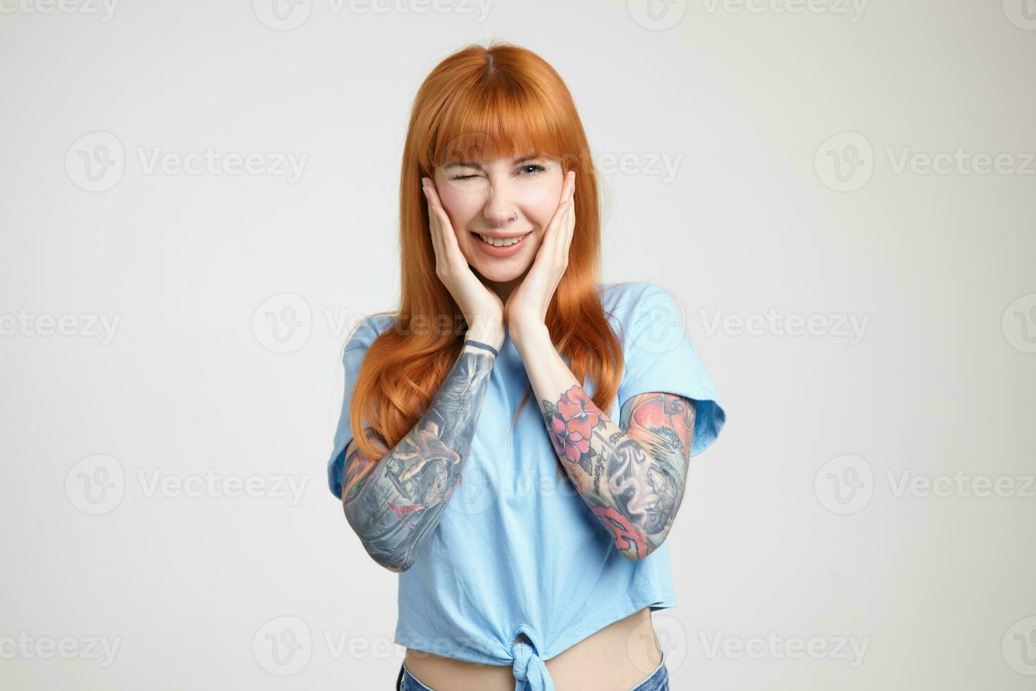Cheerful young lovely redhead tattooed female holding her face with raised hands while giving gladly wink at camera, standing against white background photo