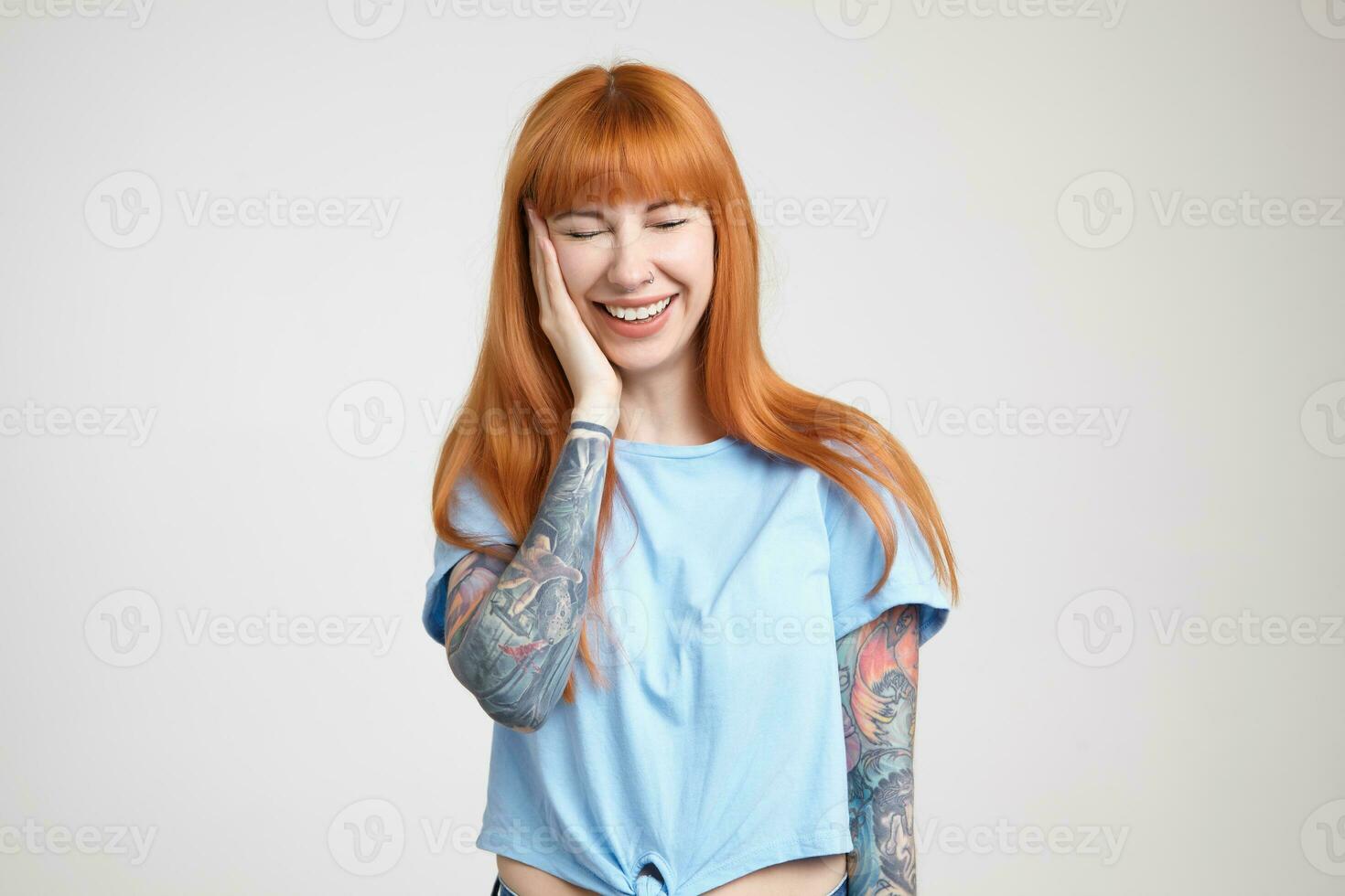 Pleasant looking young pretty long haired redhead woman holding palm on her cheek while laughing happily with closed eyes, isolated over white background photo