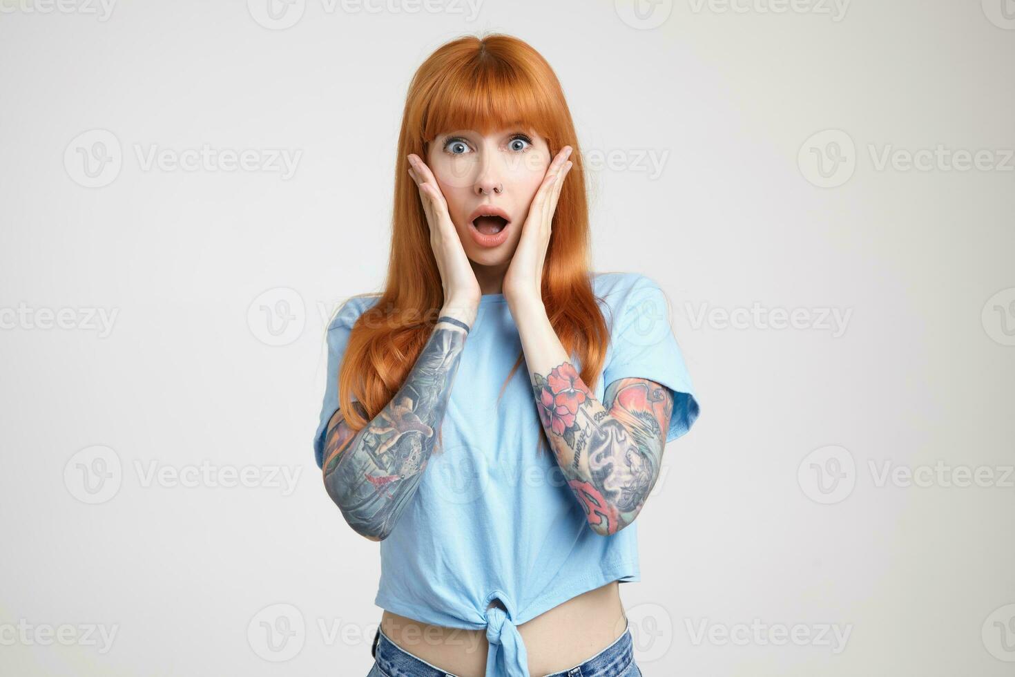 Shocked young pretty long haired redhead lady holding palms on her cheeks while looking amazedly at camera with opened mouth, isolated over white background photo
